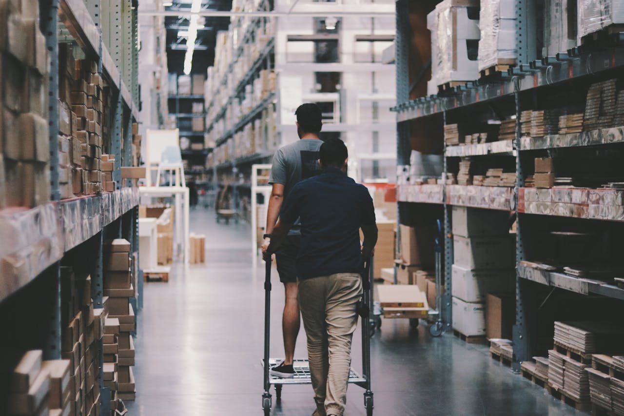 cta-02 Two men maneuver a trolley in a large warehouse filled with boxes and shelves.